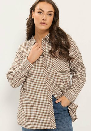 Donna con capelli lunghi e ondulati che indossa una camicia marrone e bianca a quadri larga e jeans blu, posando con una mano vicino al colletto e l'altra in tasca.
