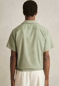 Camisa de mangas cortas de tela verde clara, con collar extendido y corte recto, de textura suave y sin patrones visibles.