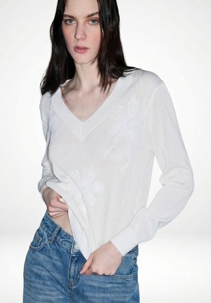 Junge Frau mit dunklen Haaren, die einen weißen V-Ausschnitt-Pullover mit dezenten Palmbaum-Mustern und blauen Jeans trägt und vor einem einfarbigen Hintergrund steht.