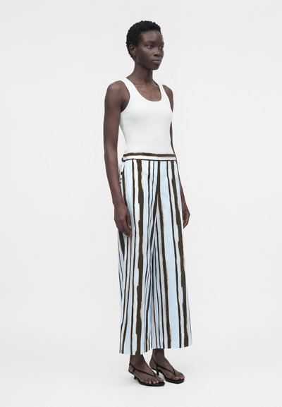 Γυναίκα που φοράει λευκό αμάνικο τοπ, φαρδιά παντελόνια με κάθετες μαύρες και ανοιχτομπλε ρίγες και μαύρα σανδάλια με ανοιχτά δάχτυλα, στέκεται ακίνητη.