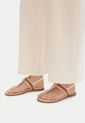 Piedi che indossano sandali bassi beige e marroni con cinturini alla caviglia e fibbia decorativa, abbinati a pantaloni crema a gamba larga.