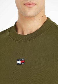Sudadera de cuello redondo en verde oliva, hecha de algodón, con un parche negro que tiene franjas rojas, blancas y azules que dice "TOMMY JEANS."