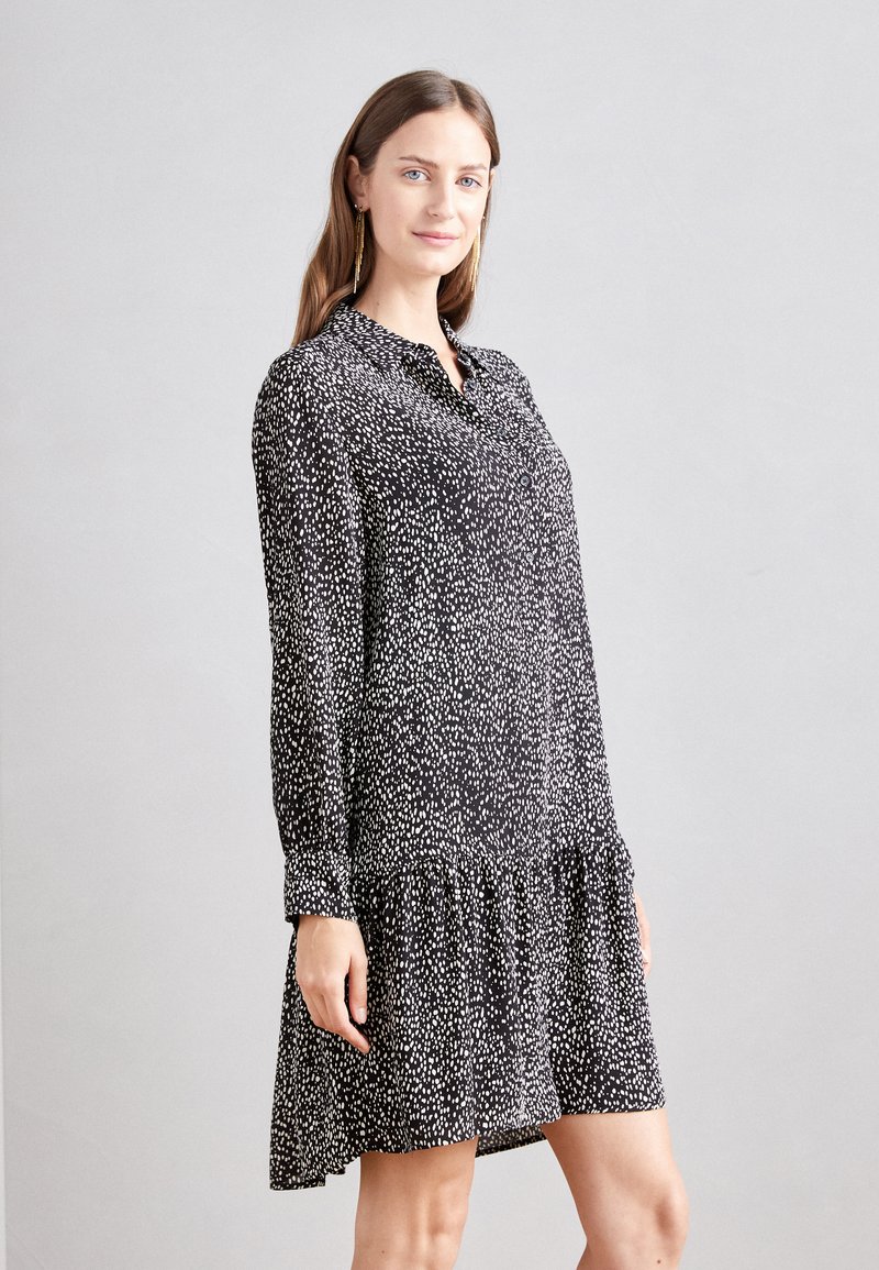 Čierno-biel dress s bodkami, dlhými rukávmi, so zapínaním na gombíky na prednej strane a riaseným lemom. Vyrobené z ľahkej tkaniny s hladkou textúrou.