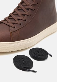 Brown leather sneaker with a high-top design, textured surface, cream rubber sole, and two black flat laces beside it.