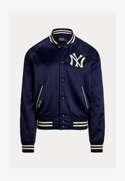 Tengerészkék szatén bomberdzseki fehér csíkos gallérral, mandzsettákkal és szegéllyel, New York Yankees logóval a mellkason, elején gombos rögzítéssel.