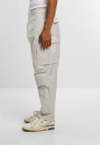Pantaloni cargo grigio chiaro con tasche laterali, gambe affusolate e una texture liscia, abbinati a sneakers beige con dettagli neri.