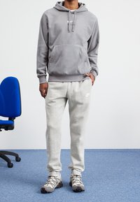 Felpa grigia in pile con tasca frontale e lacci, abbinata a pantaloni della tuta grigi chiari con un logo bianco, completata da sneakers grigie.