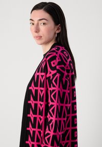Melna kardigāna ar izteiktu rozā ģeometrisku rakstu, adīta tekstūra un brīva piegriezuma, ko valkā persona ar taisniem matiem.