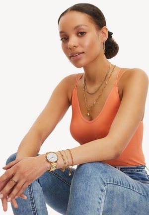 Top a righe arancione, gioielli dorati e un orologio d'argento. La modella ha i capelli mossi e si siede in modo informale con le mani appoggiate sulle ginocchia. Visibili dei jeans di denim.