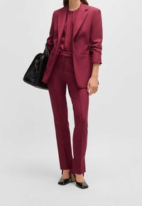 Kobieta w bordo dopasowanym garniturze z dopasowaną bluzką, czarną torebką i czarnymi butami na wysokim obcasie na jasnym, jednolitym tle.