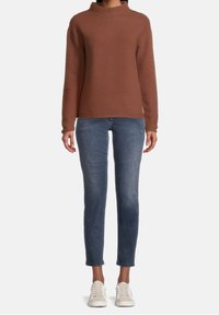 Brun ribbad tröja med hög krage, tillsammans med smala blå jeans och ljusa sneakers, stående på en enkel bakgrund.