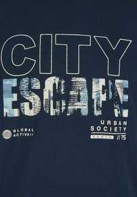 Marineblauwe katoenen t-shirt met grote witte letters "CITY ESCAPE," voorzien van blauwe fotografische patronen in de tekst en kleine merksymbolen eronder.