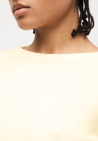 Gros plan sur le bas du visage, le cou et l'épaule d'une personne portant un pull en maille jaune clair, avec des cheveux tressés visibles.