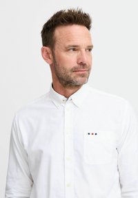 Fehér pamut ing, zsebes kivitelben. A zseben kis színes csíkok találhatók, és gombolható gallérral rendelkezik.