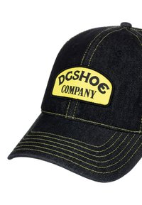 Svart denimkeps med gul sömnad och en gul etikett med texten "DCSHOE COMPANY". Kurvad skärm med sexpanelig design.