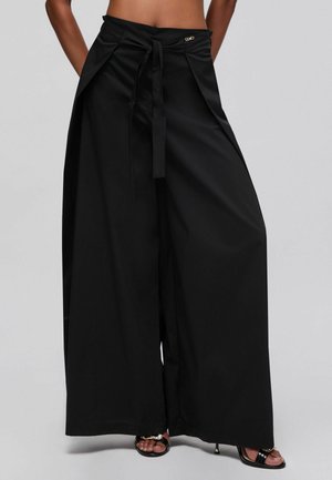 Donna che indossa pantaloni neri a vita alta e gamba larga con cintura da annodare e tacchi neri aperti davanti a uno sfondo grigio semplice.