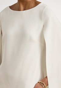 Šviesiai gelsvas viršus su plačia apvaliomis iškirpte ir skraistės stiliaus rankovėmis, pagamintas iš lygios, laisvai krentančios medžiagos. Turi subtilią tekstūrą ir gražią kritimą.