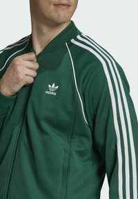 Grön zip-jacka i mjukt tyg, med vita tre-strimmor på axlarna och en liten broderad logotyp på bröstet.