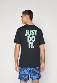 Sort bomulds t-shirt med korte ærmer og rund halsudskæring, med grøn "JUST DO IT." tekst på ryggen. Kombineret med blå mønstrede shorts.
