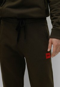 Dunkelolivgrüne Jogginghose mit Kordelzugbund, mit einem kleinen roten rechteckigen Etikett mit "HUGO" auf der linken Tasche versehen.