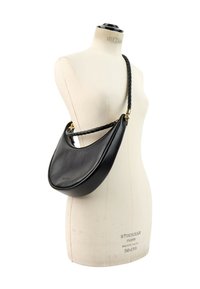 Sac d’épaule en cuir noir en forme de croissant avec une bandoulière tressée exposé sur un mannequin en forme de robe blanche.