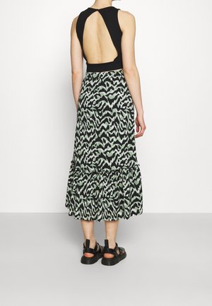 Frau trägt ein ärmelloses schwarzes Oberteil mit offenem Rücken und einen knielangen Rock mit Animal-Print, kombiniert mit schwarzen Sandalen, steht vor einer weißen Wand.