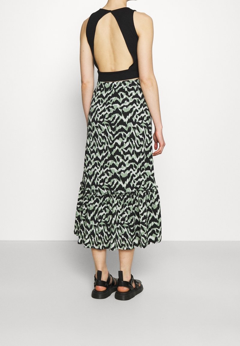 Femme portant un haut noir sans manches à dos ouvert et une jupe mi-longue à imprimé animal, associée à des sandales noires, debout contre un mur blanc.