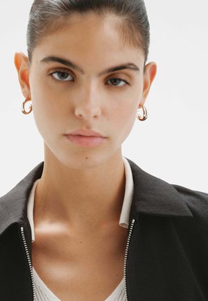 Pendientes de aro dorados, una chaqueta negra con cuello y cremallera, con una camiseta blanca texturizada debajo. Piel suave, cabello castaño medio.