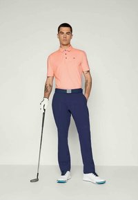 Svetloružová polo košeľa s tromi gombíkmi a logom na hrudi, v kombinácii s tmavomodrými nohavicami a bielymi golfovými topánkami, drží golfovú palicu.
