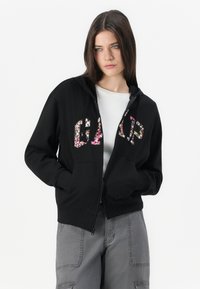 Black zip-up hoodie featuring a floral "GAP" logo on the chest, with a front pocket and a drawstring hood. Fabric appears soft and relaxed.