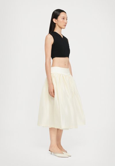 Top negro corto con bordes con forma de concha, combinado con una falda de satén blanca de talle alto y plisada. La modelo lleva mules nude con punta.