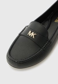 Zwarte leren loafers met een gestructureerde afwerking, voorzien van een gouden MK-logo accent aan de voorkant en decoratieve stiksels langs de randen.