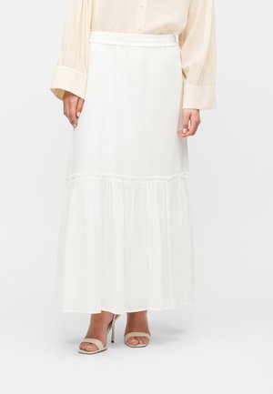 Mujer con una falda larga blanca de volantes, blusa beige con puños abotonados y sandalias beige abiertas de tacón alto, de pie.