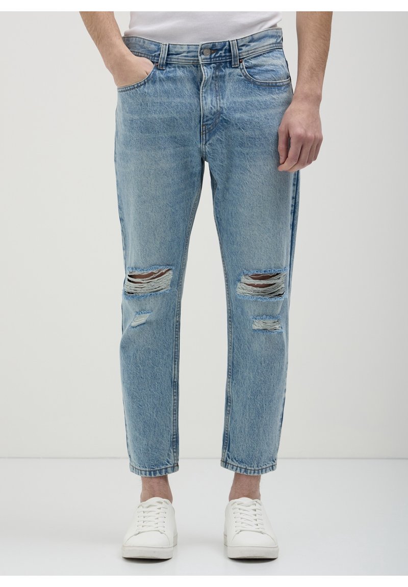 Homme portant un jean bleu clair déchiré et des baskets blanches, debout avec une main dans la poche devant un fond clair uni.