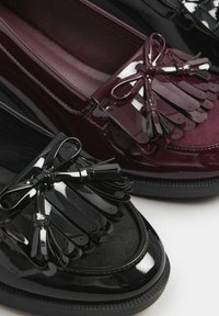 Svarta och burgundy loafers i blankt patenterat konstläder, med rosettdekorationer och fransdetaljer på framsidan. Platta sulor, texturerad yta.
