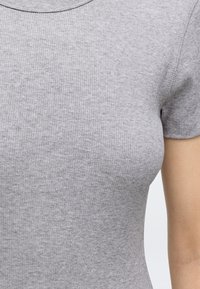 Gros plan d'une personne portant un t-shirt gris côtelé ajusté à manches courtes, montrant le torse et le haut du bras.