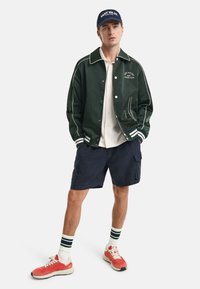 Giacca da baseball verde scuro con piping bianco, camicia bianca a bottoni, pantaloni corti blu navy, scarpe da ginnastica rosse e calzini a righe bianchi e verdi.