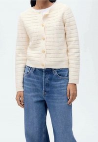 Cardigan en tricot crème avec des rayures texturées et des boutons dorés, associé à un jean en denim bleu. Le cardigan présente des manches longues et un col rond.
