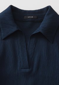 Blusa blu navy con texture e colletto, caratterizzata da piccoli dettagli a pieghe e una patta. L'etichetta riporta la scritta "OPUS" sul collo.