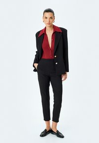 Veste noire cintrée sur une chemise rouge à boutons, assortie à un pantalon noir ajusté et des mocassins brillants. Textures lisses, design structuré.