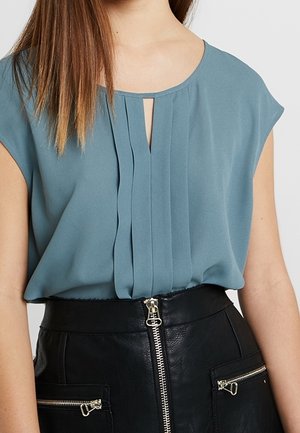 Kvinde iført en ærmeløs teal-bluse med for-pliéer, stoppet ned i et sort lædernederdel med lynlås foran og lynlåslommer.