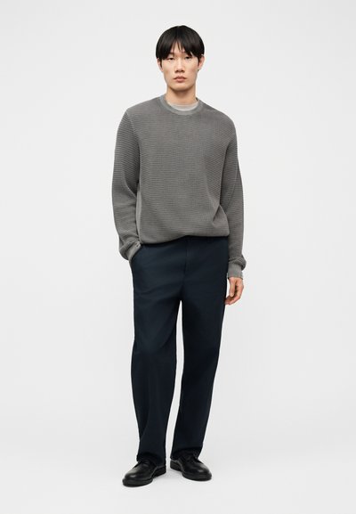 Homme portant un pull gris texturé à manches longues, un pantalon bleu marine foncé et des chaussures noires, debout les mains dans les poches devant un fond uni.