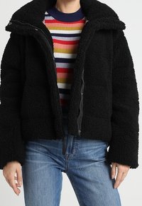 Sort fleecejakke med en struktureret overflade, høj krave og lynlås, lagd over en flerfarvet stribet sweater.