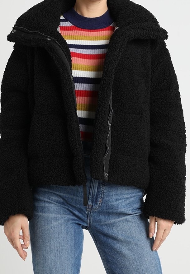 Sort fleecejakke med en struktureret overflade, høj krave og lynlås, lagd over en flerfarvet stribet sweater.