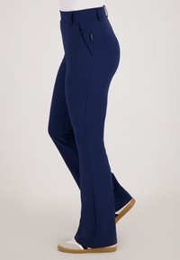 Slim fit donkerblauwe broek met zijzakken, gedragen met witte en beige sneakers door een persoon tegen een effen achtergrond.