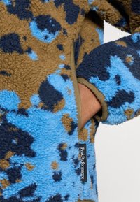 Fleecejack met een blauw, bruin en donkerblauw gespikkeld patroon; heeft een groene rand en een zijzak. Logo-label aan de onderkant.