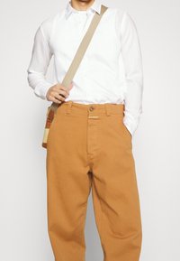 Vit långärmad skjorta med knäppt krage, tan bomullsbyxor med framfickor och en beige axelväska med bruna accenter.