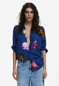 Blue floral button-up shirt with short sleeves, featuring vibrant multicolored flower patterns and a relaxed fit. Paired with light blue denim.