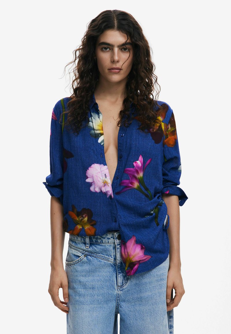 Blue floral button-up shirt with short sleeves, featuring vibrant multicolored flower patterns and a relaxed fit. Paired with light blue denim.