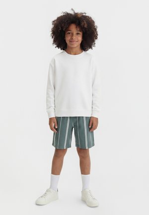 Dziecko o kręconych włosach, ubrane w białą bluzkę z długim rękawem, zielono-białe pasiasty szorty, białe skarpetki i białe tenisówki, stojące i uśmiechające się.